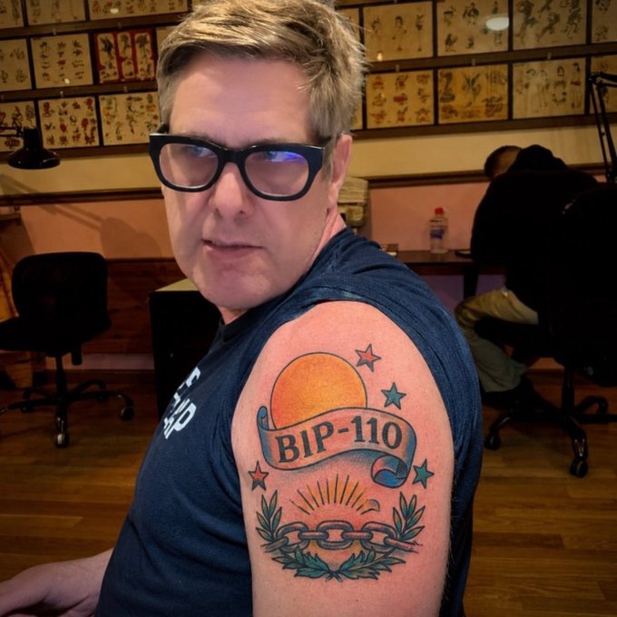 A man with glasses poses to show off his new traditional-style tattoo featuring a banner that reads "BIP-110" above a sunrise and broken chain design, visible on his shoulder in what appears to be a tattoo parlor with flash sheets on the walls. The post's caption suggests "Fred" recently got this tattoo to commemorate BIP-110, a Bitcoin Improvement Proposal, humorously implying this short-lived dedication might soon shift to another topic.