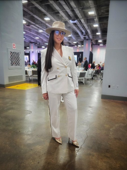 A woman in a white pinstripe suit, gold metallic heels, and a tan cowboy hat poses for a photo in an event space.
