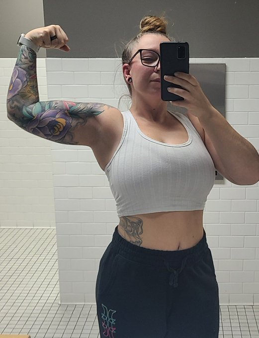 A mirror selfie of a woman with blonde hair in a bun, glasses, and a colorful floral sleeve tattoo, wearing a white crop top and dark sweatpants in a white-tiled bathroom.