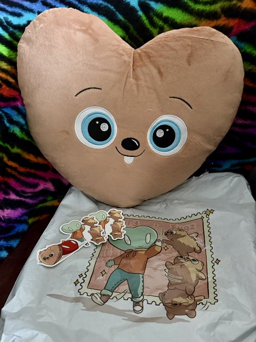 A heart-shaped plush toy with a cute face sits next to a t-shirt featuring cartoon characters on a stamp design, placed against a colorful zebra-print background.