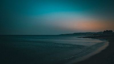 A dark, low-light photograph of a beach at dusk or night with the ocean on the left and a faint horizon showing distant lights.