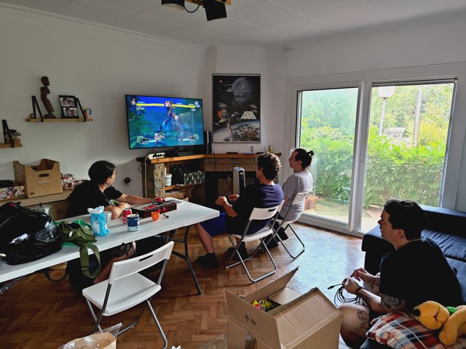 A group of five people are gathered in a living room playing a video game on a wall-mounted TV displaying what appears to be a Super Smash Bros. battle, with cardboard moving boxes scattered around the room. The casual setting and team hashtag suggest this is a fun team bonding moment for the Arkham crew, likely during a move-in or break time.