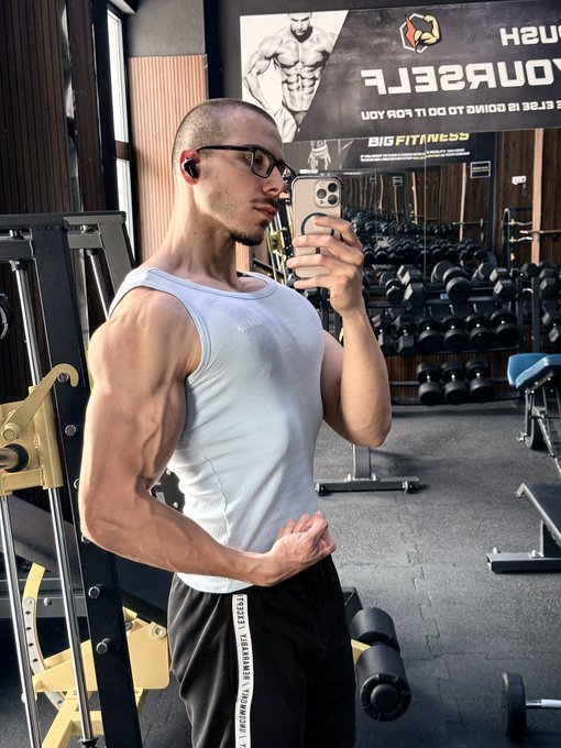 A muscular man in a white tank top takes a mirror selfie at Big Fitness gym, flexing his biceps prominently. The image, paired with the caption "Average $STONK holder," humorously plays on the meme suggesting that fitness gains are a direct result of stock market profits.