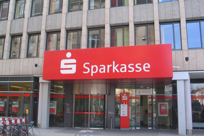 A Sparkasse bank branch with a large red “Sparkasse” sign and storefront entrance. The image illustrates the accompanying post’s claim that the German bank is integrating crypto trading, including BTC, ETH, XRP, SOL, and POL, into its banking app.