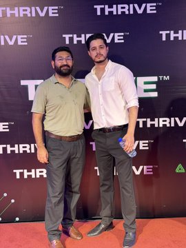 Two men pose at the launch of "THRIVE" (Thrive Web3) in Bahria Town Islamabad, set against a branded backdrop repeating the THRIVE™ logo, with one attendee holding what appears to be a water bottle on the red carpet, capturing the celebratory atmosphere of the platform's debut focused on free crypto, AI, and digital skills education.