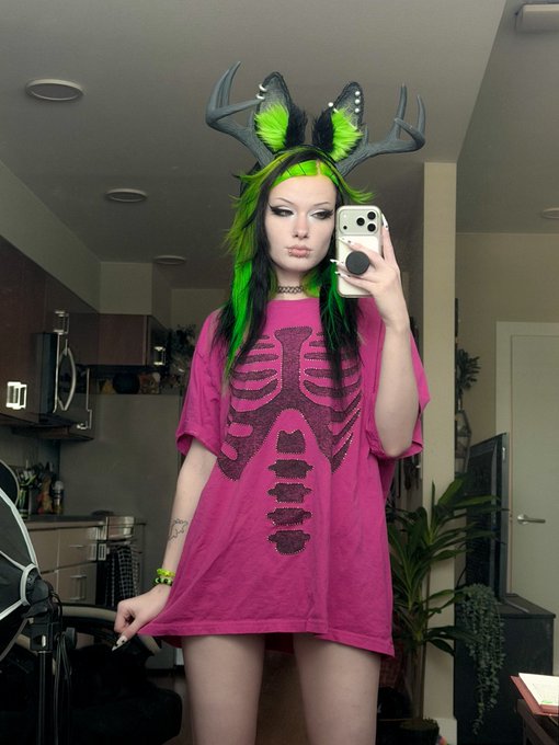 A person takes a mirror selfie wearing a pink shirt with a black ribcage/skeleton design and a green-and-black wig with antler ears, styled after a jackalope to match the "Jackalope b!tch" caption. Their gothic makeup and bold accessories complete the alternative aesthetic.