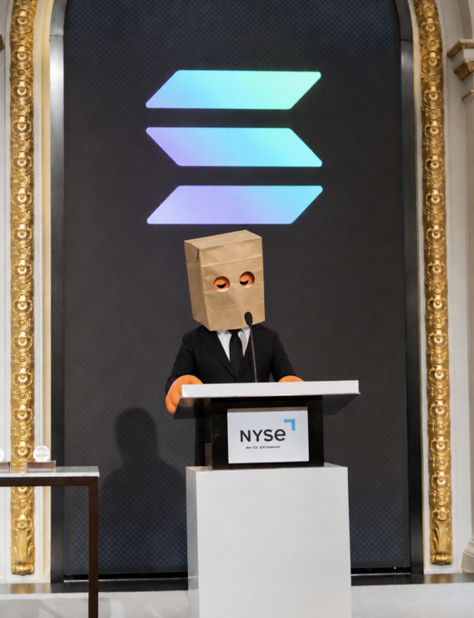 A figure wearing a suit and a brown paper bag with eye holes stands at the NYSE podium under a Solana logo. The "bag stays on" caption reinforces the anonymous, meme-inspired persona concealing their identity while addressing the iconic New York Stock Exchange.