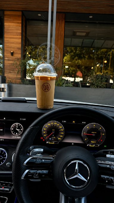 The interior dashboard of a Mercedes-Benz car featuring an iced coffee in a clear plastic cup with a 'W' logo sitting on the dash near the windshield.