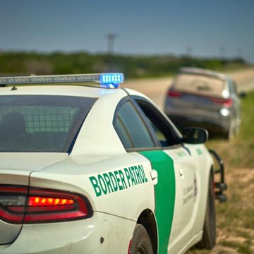 A Border Patrol police car with its light bar on, stopped on the side of a road or highway shoulder with another vehicle visible in the distance under a clear sky.