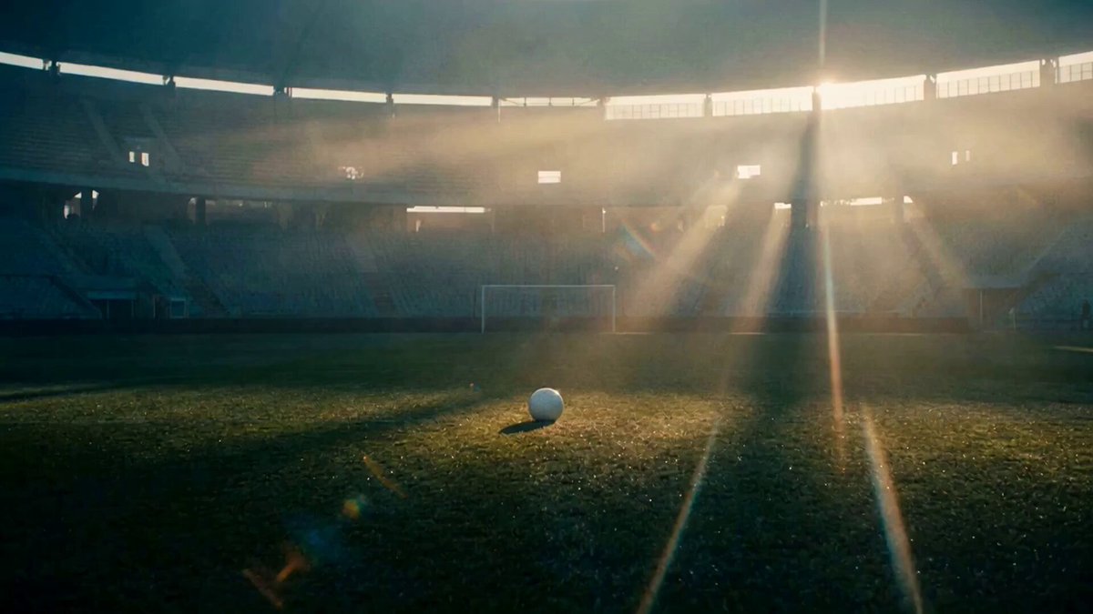 The image features an empty soccer field with a white ball sitting in the center. The sun is shining brightly through the stadium, creating a beautiful glow around the scene. The photo appears to be taken from inside the stadium or at a close-up view of the ball on the field.