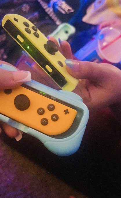 A close-up photo of a person's hands holding yellow Nintendo Switch Joy-Con controllers inserted into light blue wrist straps, with a Sonic the Hedgehog item visible in the background.