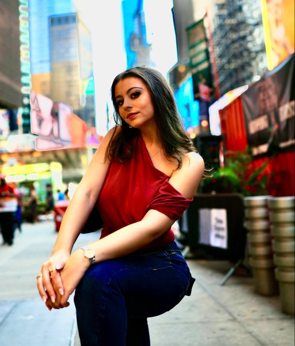 A young woman with long dark hair, wearing a red off-the-shoulder top and blue jeans, posing on a vibrant city street resembling Times Square.