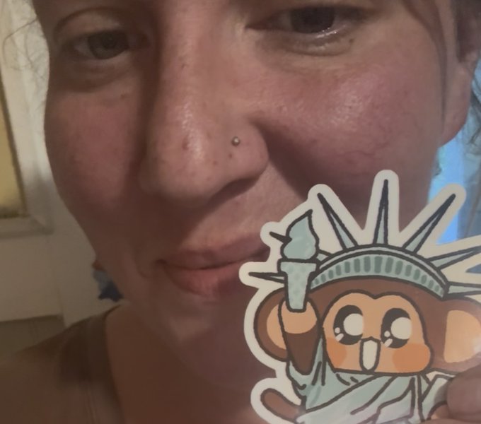 A close-up selfie of a person's face holding up a cartoon sticker depicting a monkey dressed as the Statue of Liberty holding a torch.
