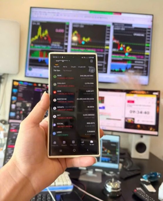 A hand holds a smartphone displaying a crypto portfolio app showing major altcoins like PEPE, SHIB, BABYDOGE, SERAPH, and NOT with notable 24-hour gains, while a multi-monitor trading setup with candlestick charts and a timer reading "09:34:48" fills the background—visually capturing the FOMO-driven altcoin chase the post warns against.