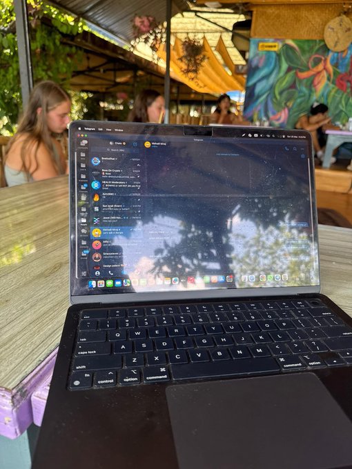 A MacBook laptop sits open on a wooden table displaying what appears to be a messaging interface on Telegram, with people working in the background of a tropical cafe setting.