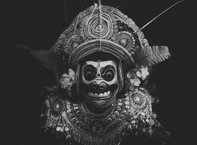 A black and white photograph of a person in elaborate traditional ceremonial attire, featuring an ornate beaded headdress, a fierce mask with prominent teeth, and decorative floral and beaded elements around the face.