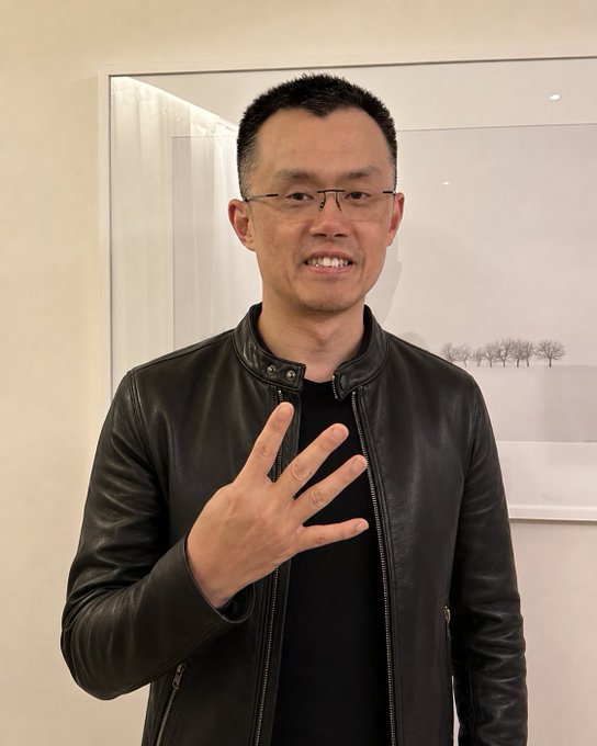 A smiling man with glasses and a black leather jacket holds up four fingers in front of a framed minimalist landscape painting, posing for a photo he hopes will catch the attention of Binance CEO CZ to engage with on social media.