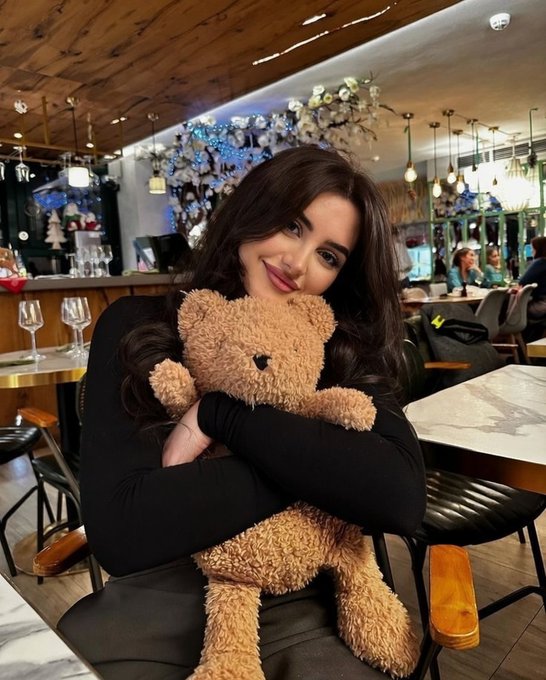 The image features a woman sitting at a table with a large teddy bear, holding it close to her chest. The photo has been captioned "Good Night friend I genuinely think the next major opportunity wave could come from ecosystems like @base & @Polymarket". This suggests that the woman might be interested in or discussing topics related to the content of the image and its connection to the post.