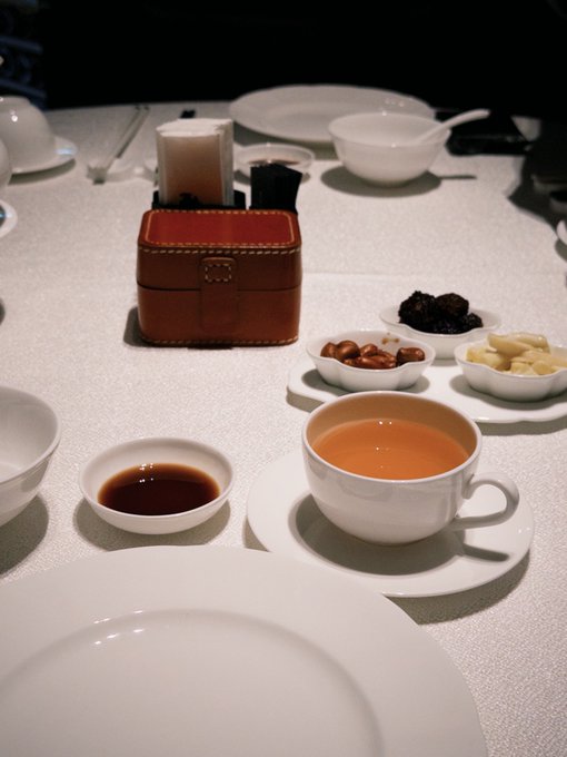 A dimly lit table setting featuring a cup of milk tea on a saucer, small dishes containing dates, nuts, and what appears to be candies or chocolates, alongside small dishes of soy sauce or vinegar, with empty plates and bowls set for a meal.