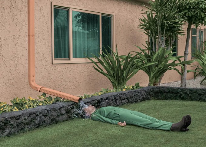 A person wearing green coveralls lies face-down on a lawn while water gushes from a cracked orange downspout onto their head, with a stucco house and tropical plants in the background.