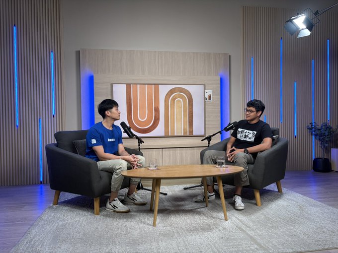 Two men sit across from each other in gray armchairs with a wooden coffee table between them, each speaking into a microphone in a podcast studio setting with a large "Un" (Base) logo screen behind them. This setup captures the interview moment described, where Nibel sits down with GitLaw and KevinCodex at the Base x NewCampus Circuit Accelerator event in Singapore to discuss their projects.
