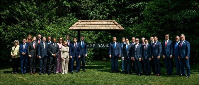 A group of world leaders and officials pose for a formal group photo outdoors at Camp David, with the "CAMP DAVID" sign visible on the gatehouse behind them, appearing to be gathered for a summit. Given the context of the "Clarity Act," the image likely depicts leaders assembled to discuss or support legislation aimed at clarifying policy positions, possibly on energy, transparency, or regulatory matters.