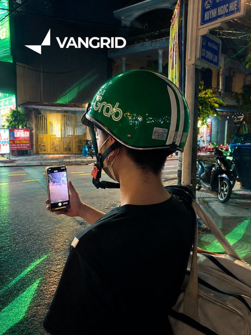 A person wearing a green Grab helmet and a black t-shirt uses a smartphone on a nighttime street, with street signs reading "HUỲNH NGỌC HUỆ" and "CHỢ ĐỒNG XUÂN" visible in the background, alongside a "VANGRID" logo in the upper left.