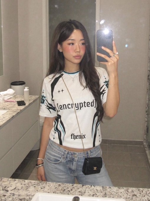 A mirror selfie of a young woman wearing a white graphic t-shirt with '[encrypted]' and 'thenix' printed on it, paired with light blue jeans and a small black crossbody bag.