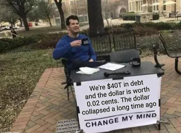 A man sits at a table outdoors with a sign reading "We're $40T in debt and the dollar is worth 0.02 cents. The dollar collapsed a long time ago. CHANGE MY MIND," holding a smaller version of the same statement.