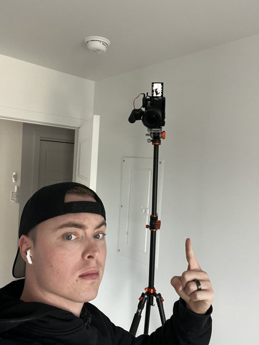 A young man wearing a black backwards cap and AirPods poses indoors, pointing upward toward a camera mounted on a tripod.