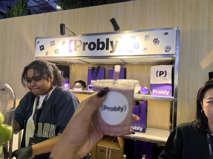 A hand holds up a small Probly-branded pouch in front of the company's illuminated booth, which displays merchandise like tote bags and keychains along with the "Probly" logo. The scene captures the casual, social atmosphere suggested by the caption's invitation to meet up and enjoy a smoothie together at the ProblyHQ pop-up.