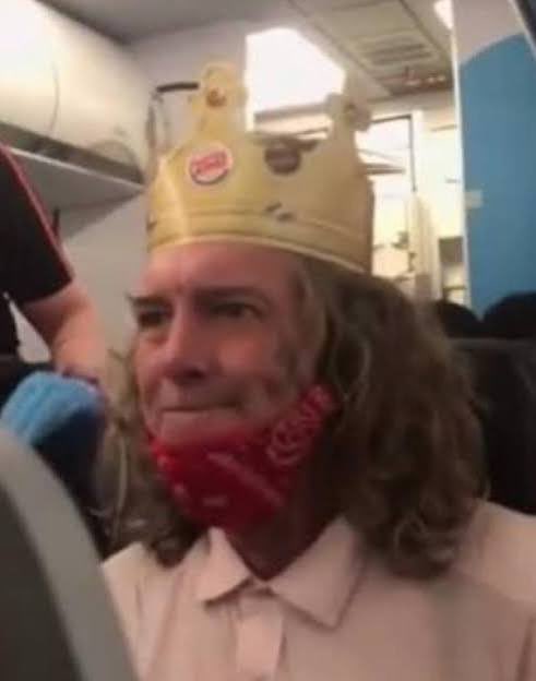 A person on an airplane wears a Burger King paper crown on their head and a red bandana over their face.