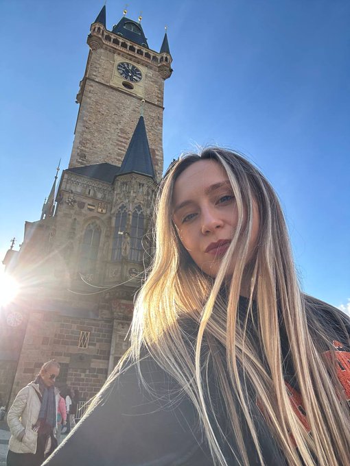 A young blonde woman takes a selfie in front of the Old Town Hall tower featuring its historic astronomical clock in Prague, likely posting a teaser the day before announcing a Web3 and Crypto Twitter meetup.