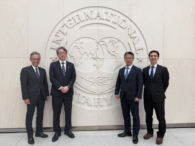 Four men in business suits pose in front of the International Monetary Fund's emblem wall, which features a world map and the visible text "INTERNATIONAL MONETARY" — apparently following a meeting related to discussions about stablecoin issuers purchasing government bonds.