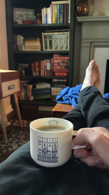 A person relaxes on a couch holding an 'ESTD' mug of coffee in front of a bookshelf filled with books.