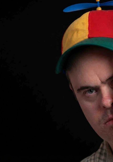 A close-up portrait of a man wearing a multi-colored propeller beanie hat with red, yellow, and green sections, set against a black background.