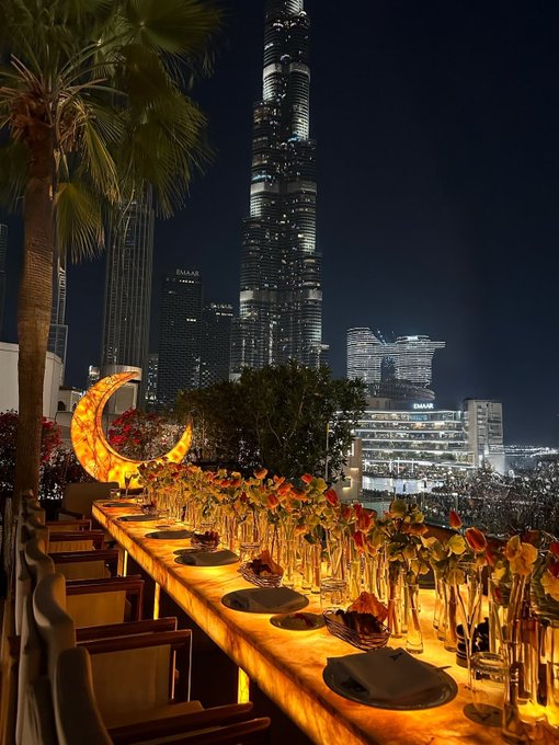 A long, glowing dining table is set for a meal on a rooftop terrace at night, with the Burj Khalifa and a building labeled 'RAKRAK' visible in the background.