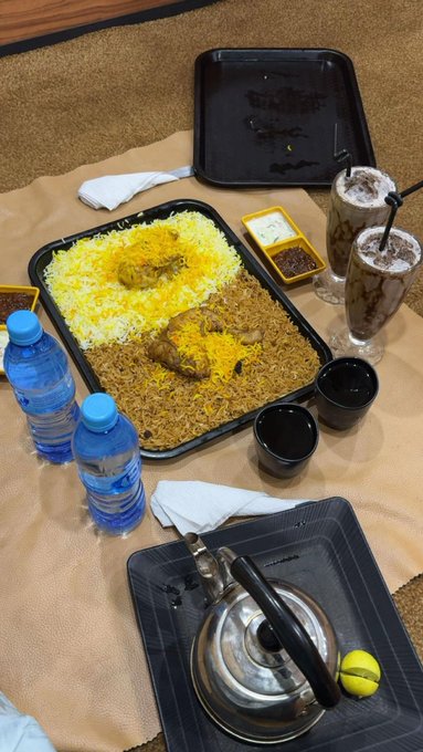 A traditional meal spread on a paper-covered table featuring a large tray of yellow and brown rice with chicken, water bottles, dark beverages, and a metal kettle.