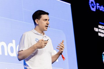 The image features a man standing on stage, wearing a white shirt with blue lettering that reads "Endo". He appears to be giving a speech or presentation while holding up his phone. The background of the image shows a large screen displaying a logo for a company named "Dash", which is likely related to the event he is speaking at. Additionally, there are two other people in the scene, one standing near the left edge and another person on the right side of the frame.
