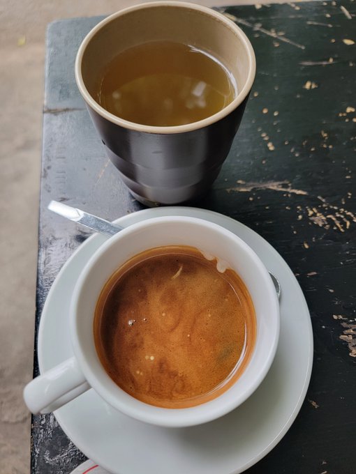 A top-down photo of two beverages on a weathered black table: a cup of green tea and a white cup of coffee with crema on a saucer.