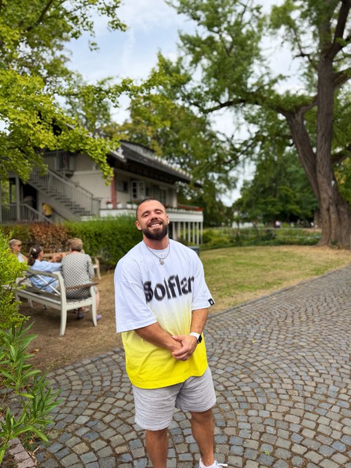 A bearded man wearing a white and yellow t-shirt with the word 'Sofler' stands smiling on a cobblestone path in a park, with people sitting on a bench in the background.