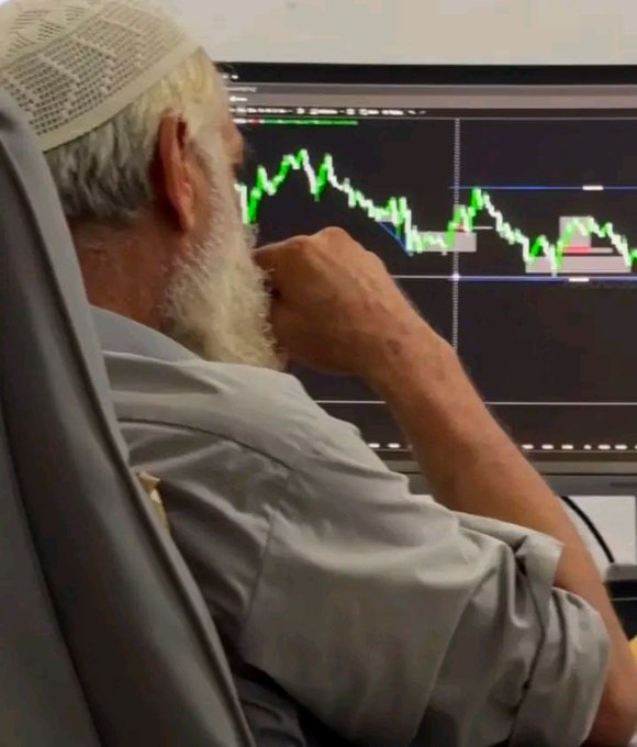 An older man with a white beard wearing a skullcap sits thoughtfully in a chair, looking at a computer monitor displaying a trading chart with green and red candlestick patterns.