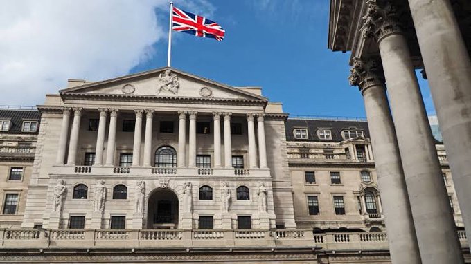 The image shows the Bank of England building with its neoclassical columned façade and Union Jack flag flying overhead. This iconic London landmark directly illustrates the post, as the Bank of England has launched a "Digital Pound Lab" to explore how a potential UK digital currency could work alongside stablecoins for cross-border trade finance.
