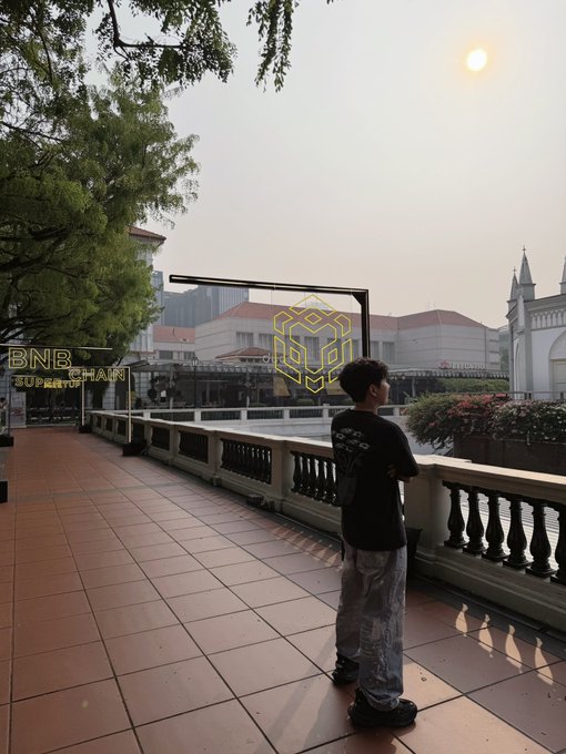 A young man stands on a balcony overlooking a cityscape, with the sun glowing brightly in the hazy sky above BNB Chain-branded signage, embodying the "insane aura" vibe of crypto culture.