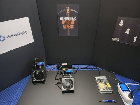 A setup with electronic devices including cameras, a multimeter, and circuit boards on a blue mat, with signs reading 'HeliumDeploy' and 'FOR HOME MINERS BY HOME MINERS' displayed on the back wall.