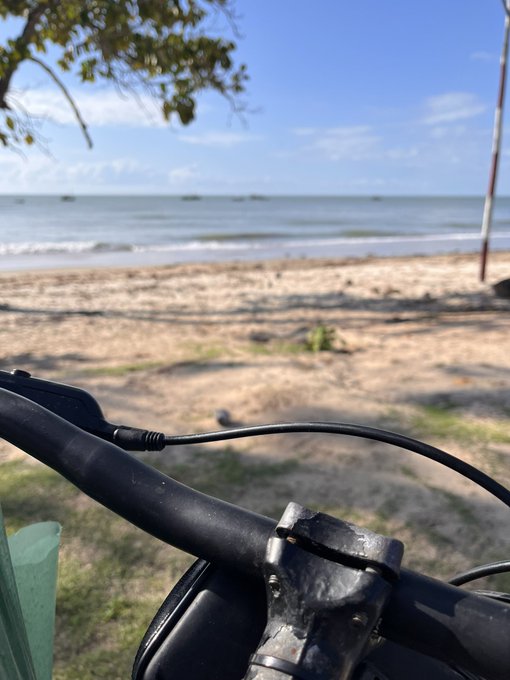 A view from behind bicycle handlebars overlooking a sandy beach with calm ocean waters and small boats visible on the horizon under a blue sky.