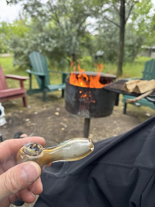 A hand holds a small glass pipe in the foreground while a metal fire pit with flames burns in the background, surrounded by patio chairs and trees in a backyard setting.