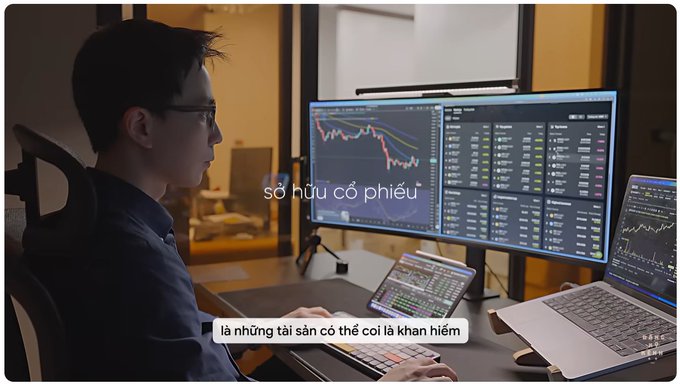 A man with glasses sitting at a desk with multiple monitors and a tablet displaying stock/crypto trading charts, with Vietnamese subtitles reading "sở hữu cổ phiếu" (owning stocks) and "là những tài sản có thể coi là khan hiếm" (are assets that can be considered scarce).