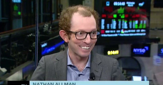 The image features a man wearing glasses and a suit talking into a microphone at a news conference or event. He appears to be discussing something related to finance or investing. The background shows a TV screen displaying stock market information, indicating that this could be an interview with Nathan Allman, who is the founder of Ondo Finance.