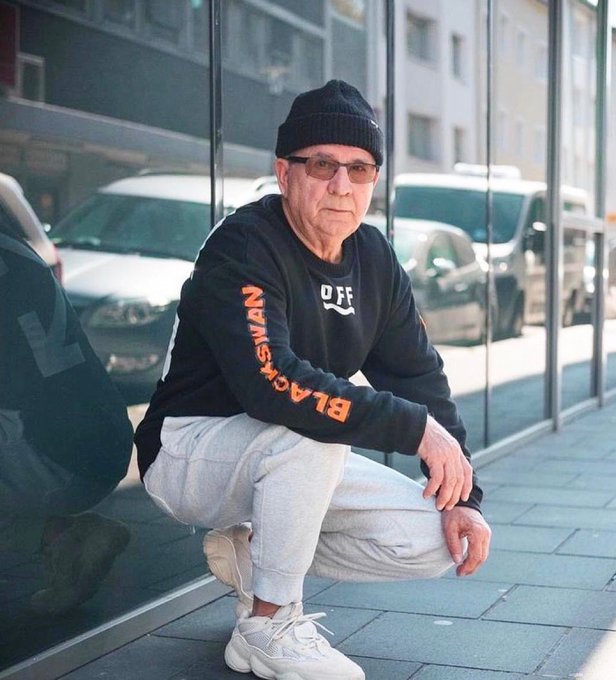 A man wearing a black beanie, glasses, and a black 'Off Black Means' sweatshirt squats on a sidewalk next to a glass storefront with parked cars visible behind him.